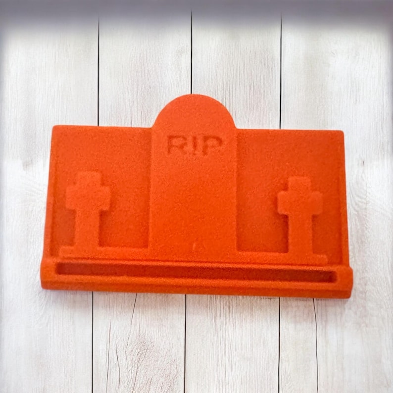 Puede incluir: Un objeto naranja en forma de l&aacute;pida con las letras "RIP" en la l&aacute;pida. Dos cruces est&aacute;n a cada lado de la l&aacute;pida. El objeto est&aacute; sobre un fondo de madera blanca.