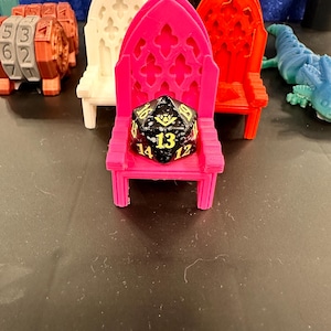 May include: A black and yellow D20 die rests on a pink miniature throne. Other colorful miniature thrones in white, green, and red are in the background. A blue lizard and a brown numerical dice are also visible.