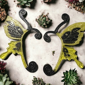 May include: Two black and yellow butterfly-shaped hair clips with intricate details. The clips are on a white background with small green plants.