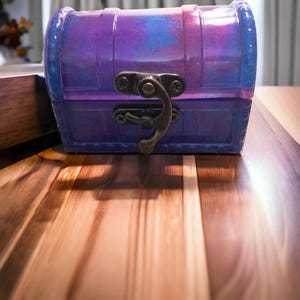 May include: A small, purple and blue treasure chest with a metal latch. The chest is sitting on a wooden surface.