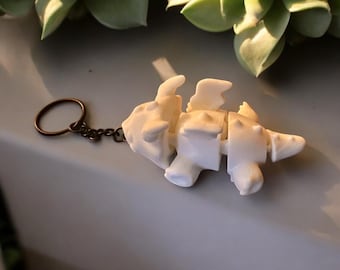Może przedstawiać: Biały, segmentowany brelok do kluczy w kształcie dinozaura z brelokiem i łańcuszkiem w kolorze brązu. Dinozaur ma małe skrzydła i kolczasty ogon. Brelok leży na szarej powierzchni, z zielonymi sukulentami w tle.