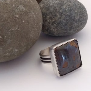 May include: A silver ring with a square, brown and black stone set in a silver bezel. The ring has a textured band with two silver lines.