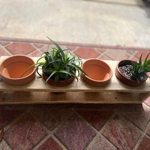 May include: A wooden planter with three terracotta pots, each containing a small green plant. The planter is made of reclaimed wood and has a rustic look.