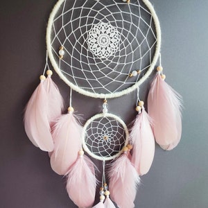 Op de afbeelding: Crèmekleurige dromenvanger met een geweven webontwerp, versierd met roze veren en houten kralen. Het woord "ANNA" is gespeld met witte kralen. Een decoratief item voor een slaapkamer.