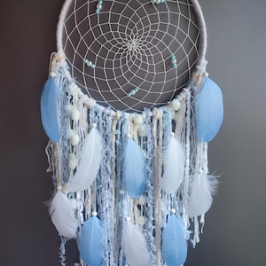 May include: A handmade dreamcatcher with a white woven web and a circular frame. The dreamcatcher features light blue and white feathers, along with blue and white yarn and small beads. The background is a solid gray.