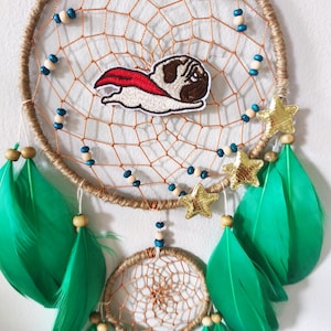 Può includere: Un acchiappasogni fatto a mano con un design a rete intrecciata, adornato con piume verdi, stelle dorate e una toppa di bulldog francese supereroe. L'acchiappasogni presenta perline blu e di legno e un secondo cerchio più piccolo.