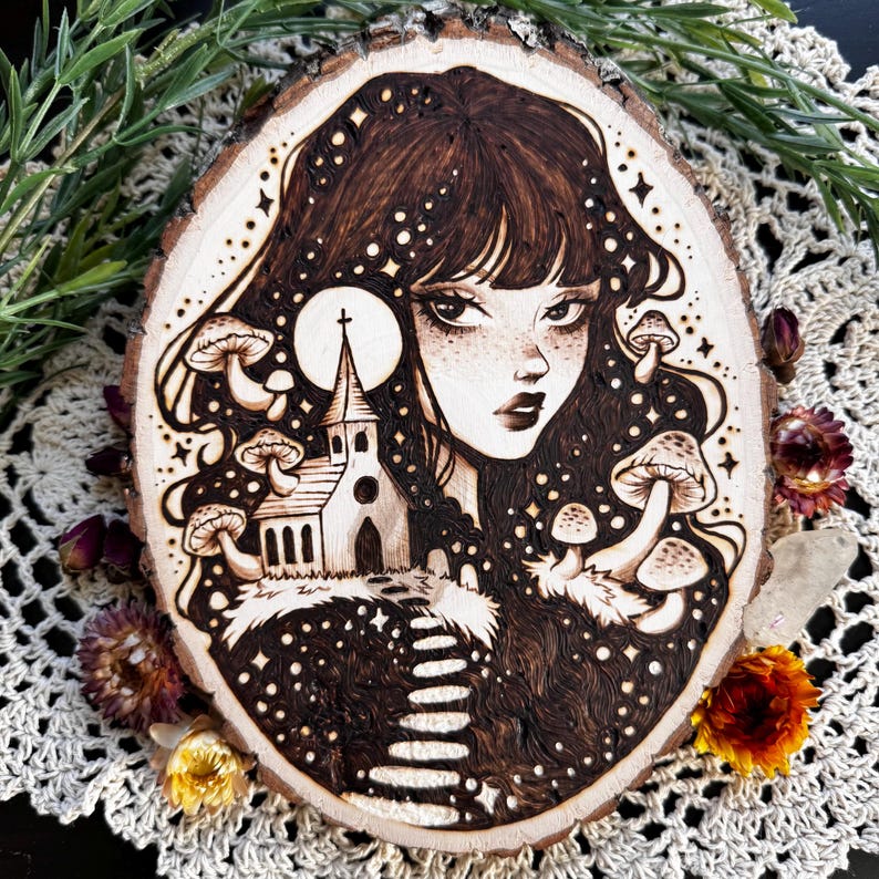Pu&ograve; includere: Una fetta di legno con un'illustrazione pirografata di una persona con lunghi capelli scuri, una piccola chiesa, una luna piena e diversi funghi. L'opera d'arte &egrave; dettagliata con ombreggiature scure e linee sottili.