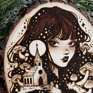 Pu&ograve; includere: Una fetta di legno circolare con un'illustrazione pirografata di una persona con lunghi capelli scuri, circondata da funghi e una piccola chiesa. Il design include accenti a forma di stella e una luna circolare dietro la chiesa.