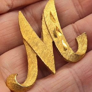 Może przedstawiać: Złota broszka z metalu w kształcie litery "M". Broszka ma teksturowaną powierzchnię i stylizowany design. Litera ma zakrzywiony, kursywny wygląd z dekoracyjnym zdobieniem na dole kształtu "J".