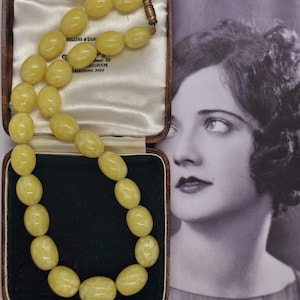 May include: A cream-coloured necklace with large, oval beads, presented in an open, brown jewellery box with a cream satin lining. The necklace is set against a black velvet interior. A vintage photograph of a woman is in the background.