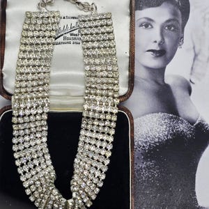 Può includere: Una collana vintage di strass con più file di cristalli trasparenti scintillanti. La collana è esposta in un portagioie foderato di velluto. Un ritratto in bianco e nero di una donna è sullo sfondo.