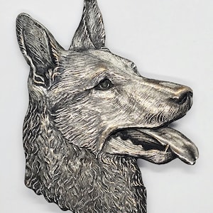 May include: A silver-colored German Shepherd dog head sculpture with its tongue out. The sculpture has detailed fur texture and is mounted on a white background. The dog's features are well-defined, with a focus on the eyes and nose.