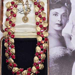 Può includere: Collana vintage color oro con strass rosso rubino e dettagli a forma di foglia, esposta in un portagioie vintage. La collana presenta un design a doppia fila con una chiusura sicura. La scatola ha un interno color crema e una base in velluto nero.