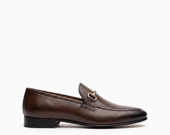 May include: A dark brown leather loafer featuring a smooth finish, a rounded toe, and a gold-toned metal horsebit detail across the vamp. The shoe has a low stacked heel and a black sole, presented in a side profile view against a plain background.
