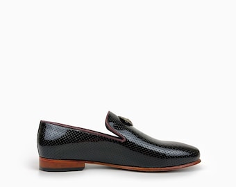 May include: A black slip-on loafer featuring a perforated leather upper with a glossy finish. The shoe has a dark maroon trim along the collar, a low stacked wooden heel, and a metallic gold-toned emblem centered on the top of the vamp.