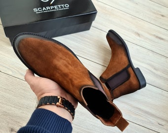 May include: A pair of brown suede Chelsea boots with elastic side panels and a black heel. The boots are being held by a hand wearing a black watch.