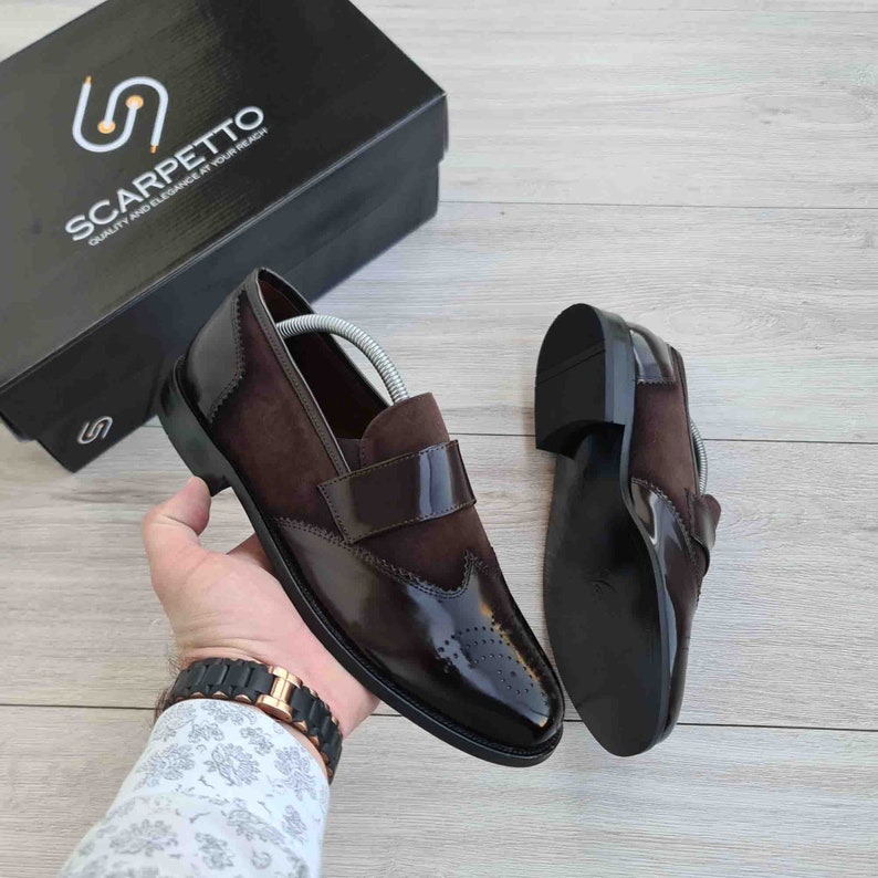 May include: A pair of brown leather slip-on shoes with a wingtip design. The shoes have a brown suede panel on the top and a black leather sole.