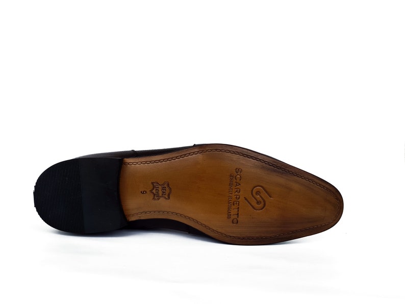 May include: The bottom of a brown leather dress shoe with a black rubber sole. The sole is stamped with the brand name "Scarpetta" and the words "Genuine Leather Made in Italy".