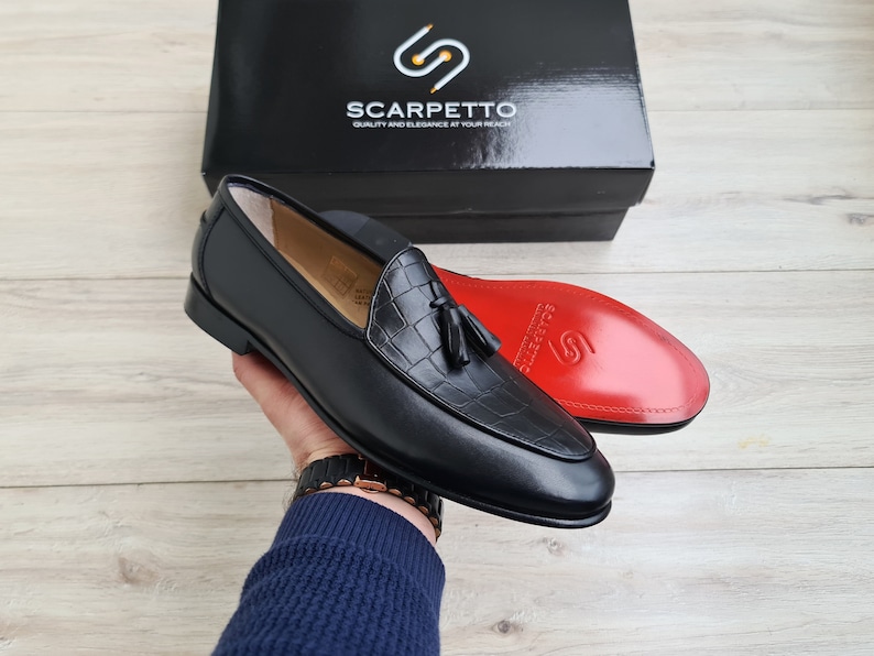May include: Black leather loafers with a tassel detail and crocodile embossed pattern. The loafers are on a person's hand and the brand name "Scarpetto" is visible on a box in the background.