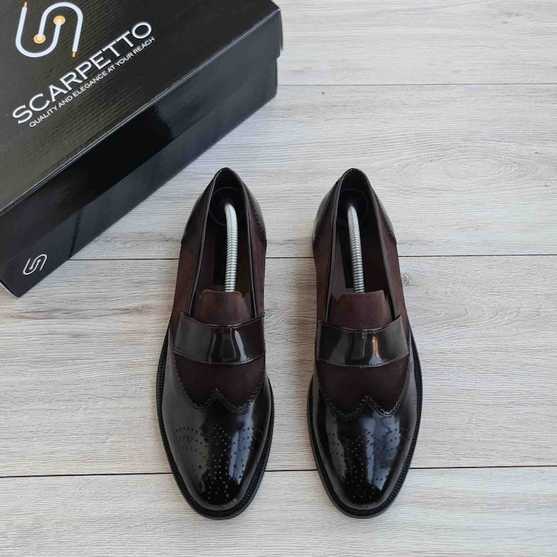 May include: A pair of brown leather loafers with a black patent leather toe cap and brogue detailing. The shoes have a suede upper and a leather sole. The shoes are on a wooden surface.