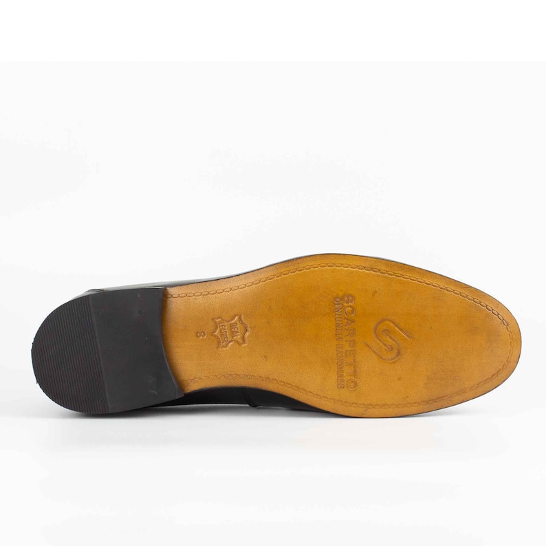 May include: Close-up of the bottom of a black leather shoe with a brown leather sole. The sole is stamped with the text "Scarpefficio" and "Genuine Leather Handmade in Italy".