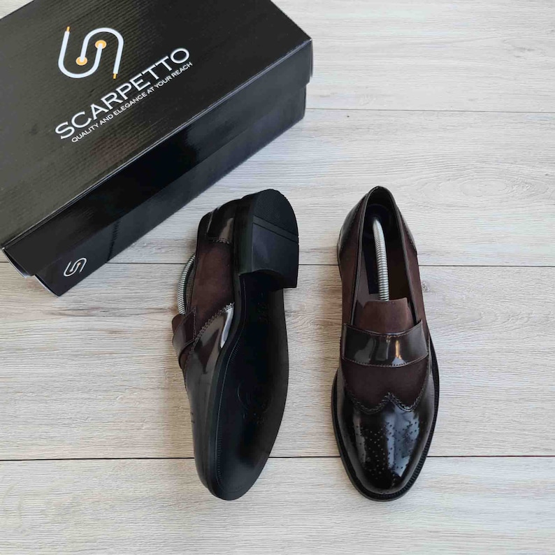 May include: A pair of brown leather loafers with a suede panel and brogue detailing. The shoes are on a wooden surface. The text "SCARPETTO QUALITY AND ELEGANCE AT YOUR REACH" is on the box.