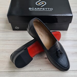 May include: A pair of black leather tassel loafers with a crocodile-embossed pattern. The loafers have a red leather lining and a black rubber sole. The shoes are displayed on a wooden surface.