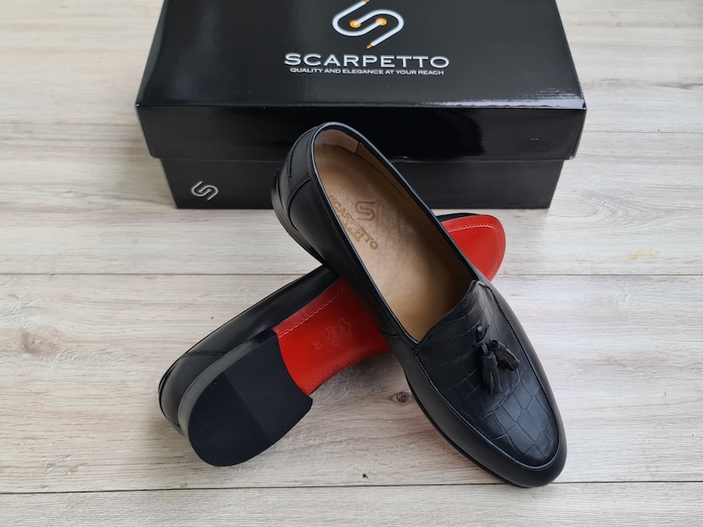 May include: A pair of black leather tassel loafers with a crocodile-embossed pattern. The loafers have a red leather lining and a black rubber sole. The shoes are displayed on a wooden surface.