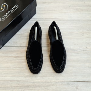 May include: A pair of black velvet loafers with a black beaded design. The loafers are on a light wood surface.