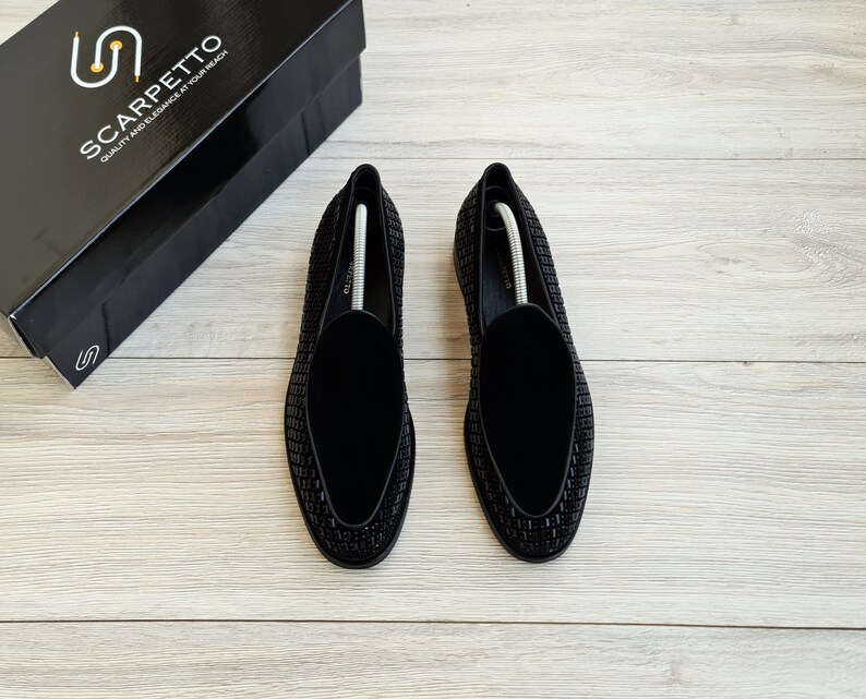 May include: A pair of black velvet loafers with a black beaded design. The loafers are on a light wood surface.