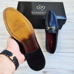 Może przedstawiać: Czarne sk&oacute;rzane loafers z ozdobną złotą klamrą. Loafers mają brązową sk&oacute;rzaną wkładkę i czarną gumową podeszwę. Buty leżą na drewnianej powierzchni.