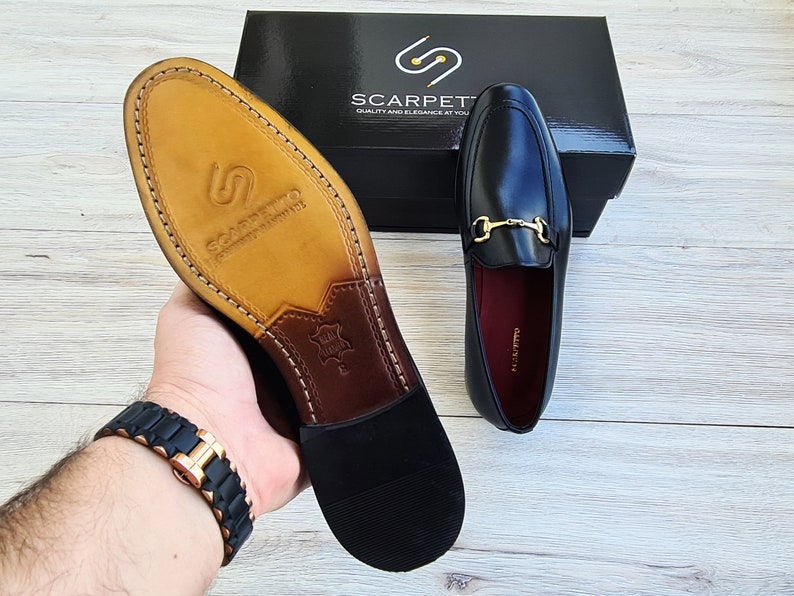 Może przedstawiać: Czarne sk&oacute;rzane loafers z ozdobną złotą klamrą. Loafers mają brązową sk&oacute;rzaną wkładkę i czarną gumową podeszwę. Buty leżą na drewnianej powierzchni.
