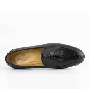 May include: Black leather tassel loafers with a crocodile embossed pattern. The loafers have a brown leather insole with the brand name "Scarpetto" printed on it.
