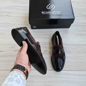 May include: A pair of brown leather loafers with a black patent leather cap toe and brogue detailing. The loafers have a slip-on design and a black rubber sole. The shoes are on a wooden surface.