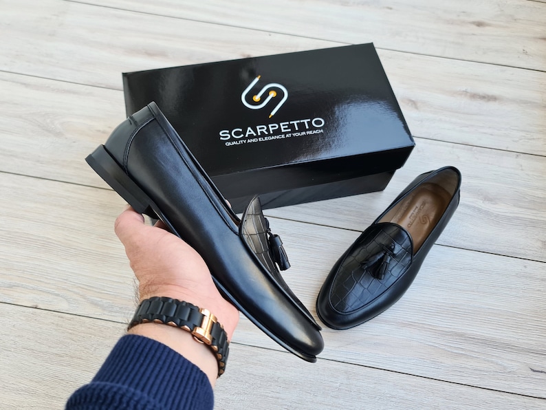 May include: Two black leather loafers with tassels. The loafers have a crocodile-embossed pattern. The loafers are on a wooden surface. The box for the loafers is in the background. The box is black with a gold logo that says "Scarpetto Quality and Elegance at your reach".