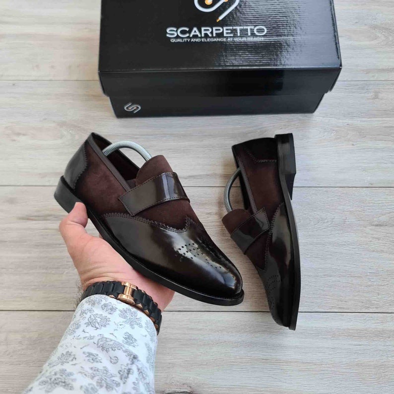May include: A pair of brown leather slip-on shoes with a suede panel and brogue detailing. The shoes are on a wooden surface.