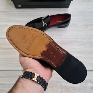May include: Close-up of the sole of a black leather loafer with a gold buckle. The sole is made of brown leather and has the text "Scarpanto" printed on it.