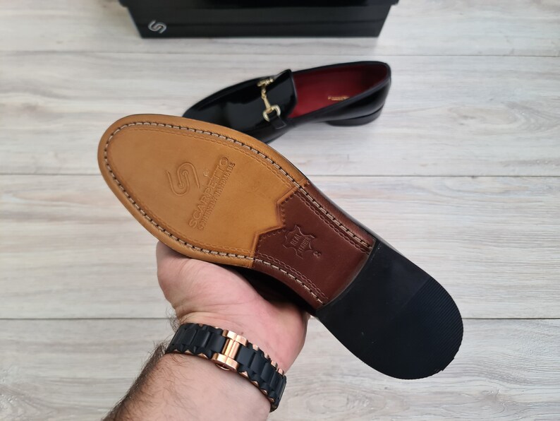 May include: Close-up of the sole of a black leather loafer with a gold buckle. The sole is made of brown leather and has the text "Scarpanto" printed on it.
