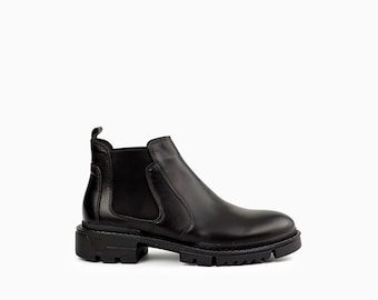 May include: A black leather Chelsea boot featuring a smooth upper, elastic side panels, a pull tab at the heel, and a thick, chunky lugged rubber sole with a deep tread pattern for traction. The boot has a rounded toe and a structured silhouette.
