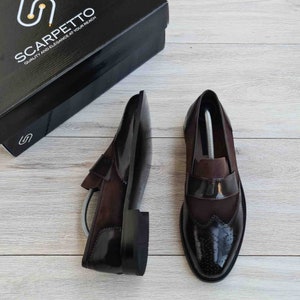 May include: A pair of brown leather loafers with a brogue design. The loafers have a black patent leather toe cap and a brown suede upper. The shoes are on a wooden surface.