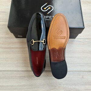 May include: A pair of black patent leather loafers with a gold metal buckle. The loafers have a brown leather sole with the brand name "Scarpetto" printed on it.