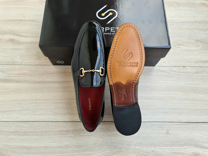 May include: A pair of black patent leather loafers with a gold metal buckle. The loafers have a brown leather sole with the brand name "Scarpetto" printed on it.