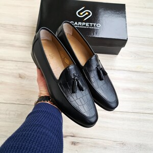 May include: A pair of black leather loafers with a crocodile embossed pattern and tassel accents. The loafers are on a wooden surface.