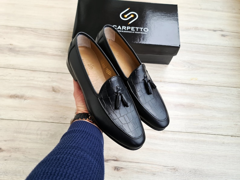 May include: A pair of black leather loafers with a crocodile embossed pattern and tassel accents. The loafers are on a wooden surface.