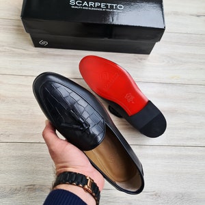 May include: A pair of black leather loafers with a tassel detail. The loafers have a red leather sole and a black leather upper. The loafers are being held in a hand.