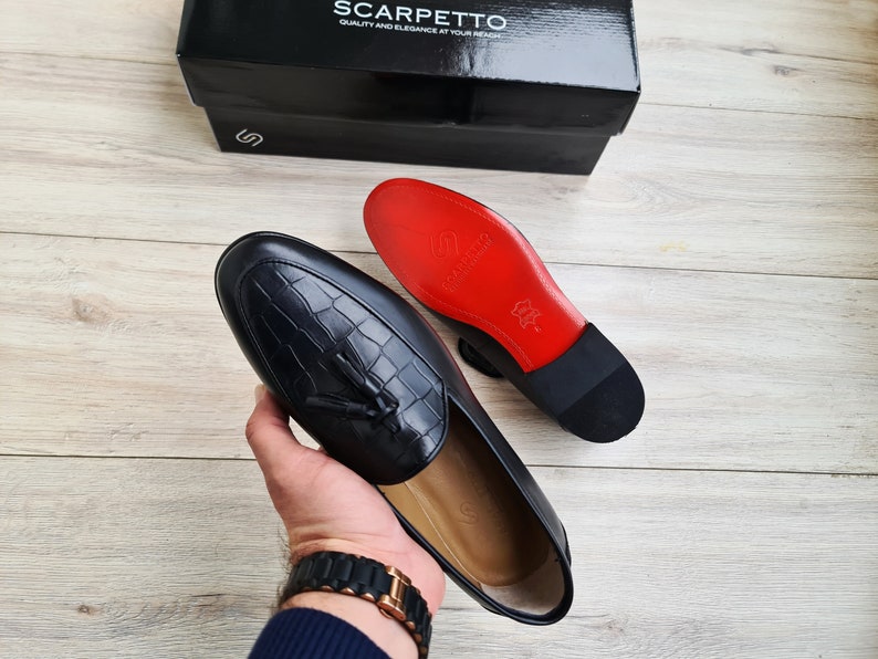May include: A pair of black leather loafers with a tassel detail. The loafers have a red leather sole and a black leather upper. The loafers are being held in a hand.