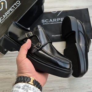 May include: Black leather monk strap shoes with silver buckles and a thick sole. The shoes are displayed with a box and a bag that read "SCARPETTO QUALITY AND ELEGANCE AT YOUR REACH".