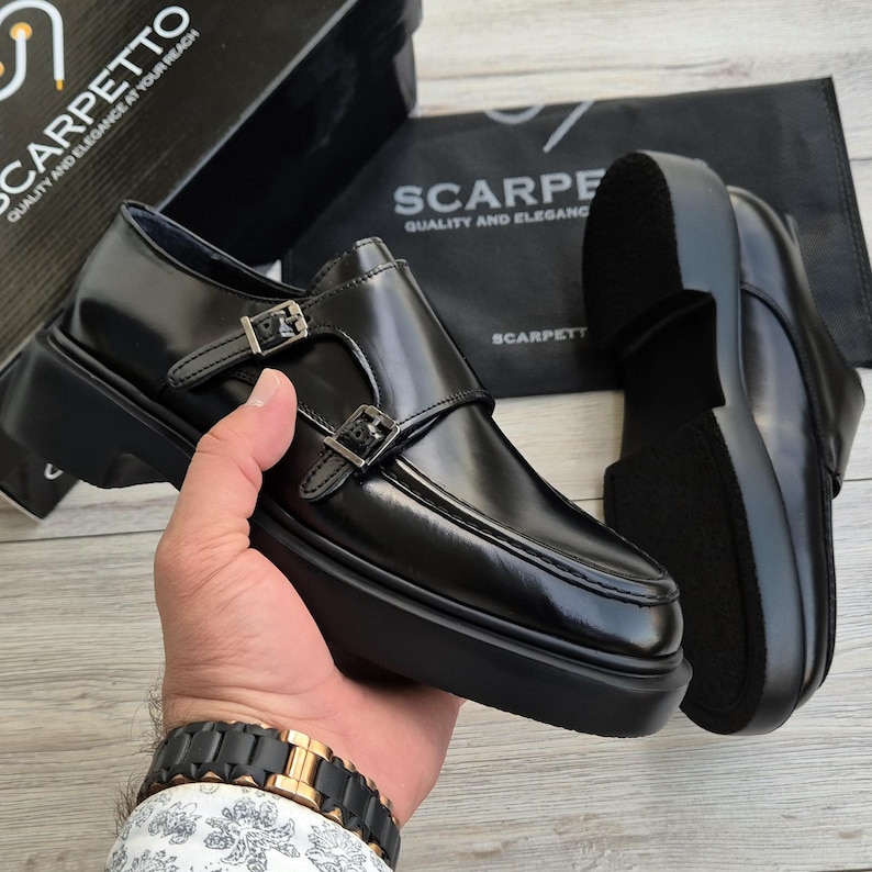 May include: Black leather monk strap shoes with silver buckles and a thick sole. The shoes are displayed with a box and a bag that read "SCARPETTO QUALITY AND ELEGANCE AT YOUR REACH".