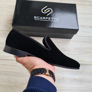 May include: Black velvet loafers with a leather sole. The loafers are being held by a hand wearing a gold watch. The brand name "SCARPETTO" is visible on a black box in the background.  Quality and Elegance at Your Reach.
