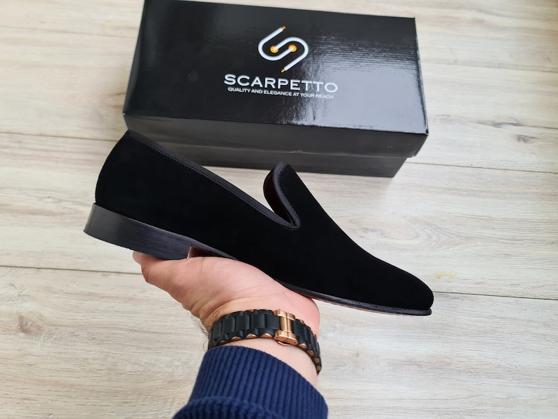 May include: Black velvet loafers with a leather sole. The loafers are being held by a hand wearing a gold watch. The brand name "SCARPETTO" is visible on a black box in the background.  Quality and Elegance at Your Reach.